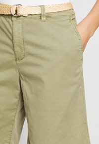 Vaaleanvihreät puuvillahousut, joissa on ryhdikäs leikkaus, sivutaskut ja kudottu beige vyö. Silkkinen rakenne näkyvissä.