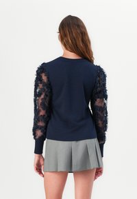 Marineblauer Pullover mit transparenten, Rüschensaum-Ärmeln. Hat einen runden Ausschnitt und gerippte Bündchen. Kombiniert mit einem grauen, gefalteten Rock.