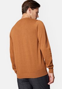 Brązowy sweter z dzianiny z prążkowanym dołem i mankietami, z okrągłym dekoltem i gładką teksturą. Pokazany z tyłu, noszony z ciemnymi spodniami w wzory.