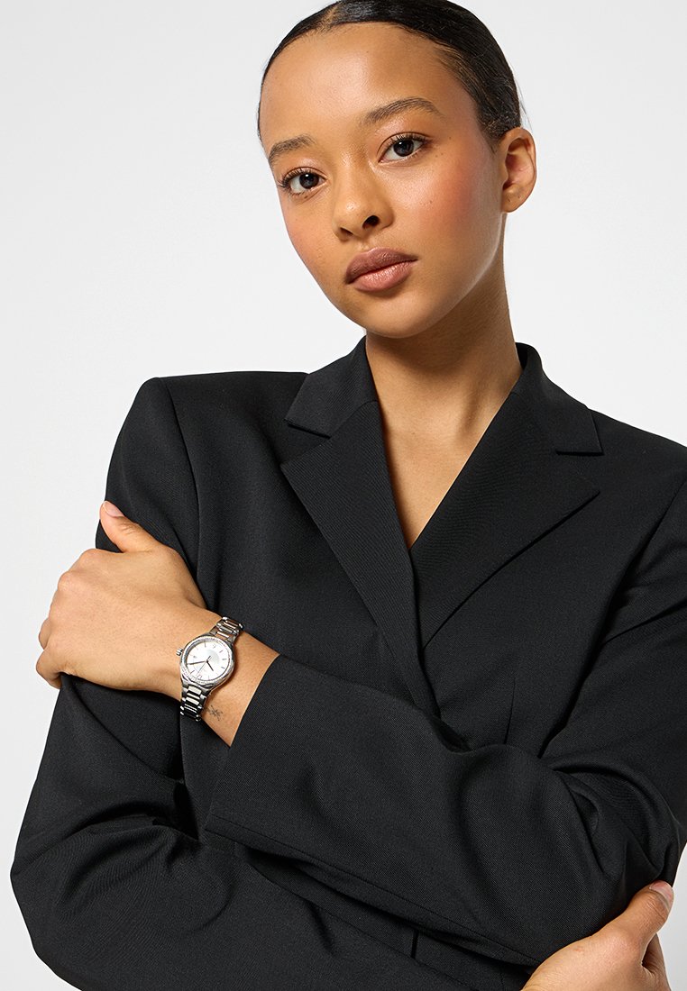 Orologio argento con quadrante bianco rotondo, bracciale in metallo a maglie, e giacca nera. Il modello posa con le braccia incrociate.