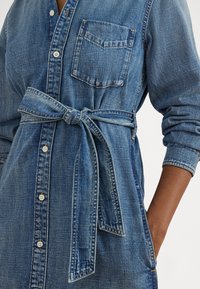 Gaiši zils džinsa krekla kleita ar pogām priekšpusē, krūšu kabatu un jostu, ko sasiež viduklī, garām piedurknēm un kabatām.