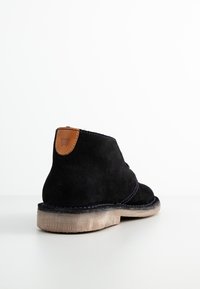 Stivaletti alla caviglia in suede nero con un pattino in pelle marrone, cuciture a contrasto e una suola in gomma chiara. Texture liscia.