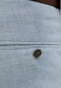 Primo piano della tasca di pantaloni in tessuto azzurro chiaro con trama, con un bottone marrone e parte di una cintura marrone visibile in alto.