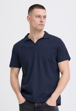 Hombre de cabello corto que lleva una camisa polo azul marino de manga corta con textura, mirando hacia la derecha con las manos en los bolsillos de unos vaqueros negros.