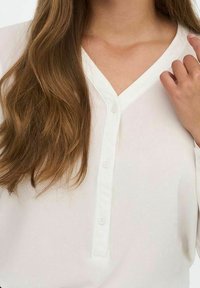 Chemisier blanc à boutons avec un décolleté en V, en tissu léger et lisse avec un léger éclat. Les boutons sont petits et blancs.