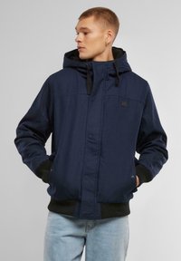 Jachetă cu glugă de culoare bleu marin, cu fermoar frontal, manșete șnur și buzunare. Are un material texturat și un logo mic pe piept.
