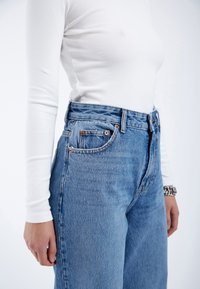High-waisted blue denim jeans with light wash and a relaxed fit, featuring copper hardware and five-pocket design. White long-sleeve top visible.