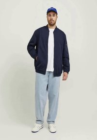 Casaco bomber azul-marinho, tecido leve, com fecho de correr à frente, bolsos laterais, combinado com uma t-shirt branca, jeans de tom azul claro e um boné azul.