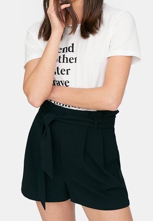 Frau trägt ein weißes T-Shirt mit Grafik, das in hoch taillierte schwarze Shorts mit gebundenem Gürtel gesteckt ist, Arme sind verschränkt und die Hand berührt ihr Gesicht.