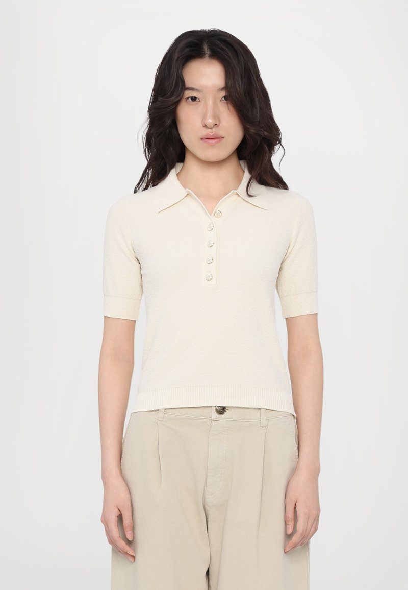 Polo à manches courtes, de couleur crème, fabriqué en tissu texturé, avec un col à boutons et cinq boutons sur le devant.