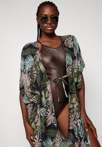 Bañador marrón con un lazo en la cintura, combinado con un kimono floral que presenta patrones de hojas verdes y beige sobre un fondo marino.