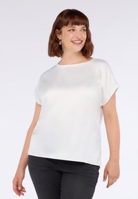 Femme aux cheveux bruns courts portant un blouse blanche à manches courtes et un pantalon noir, souriante et regardant sur le côté.