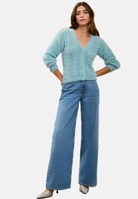 Světle modrý pletený cardigan s výstřihem do V, kabelovými vzory a knoflíky, spárovaný s širokými modrými džíny a stříbrnými botami s špičatou špičkou.