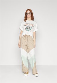 Witte grafische t-shirt met een kleurrijk arendontwerp. Lichtbeige en mintgroene chevronpatroon broek met een trekkoord in de taille. Witte sneakers.