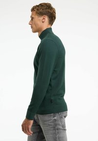 Dunkelgrüner, hochgeschlossener Pullover aus einem glatten Strickmaterial mit schmaler Passform und gerippten Bündchen. Verfügt über ein kleines Logetik am Saum.