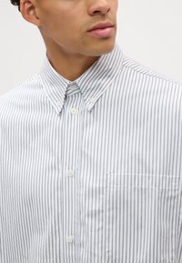Camisa blanca de botones con rayas verticales azules, cuello tipo camisa y un bolsillo en el pecho izquierdo. Fabricada en tela suave con una sutil textura.