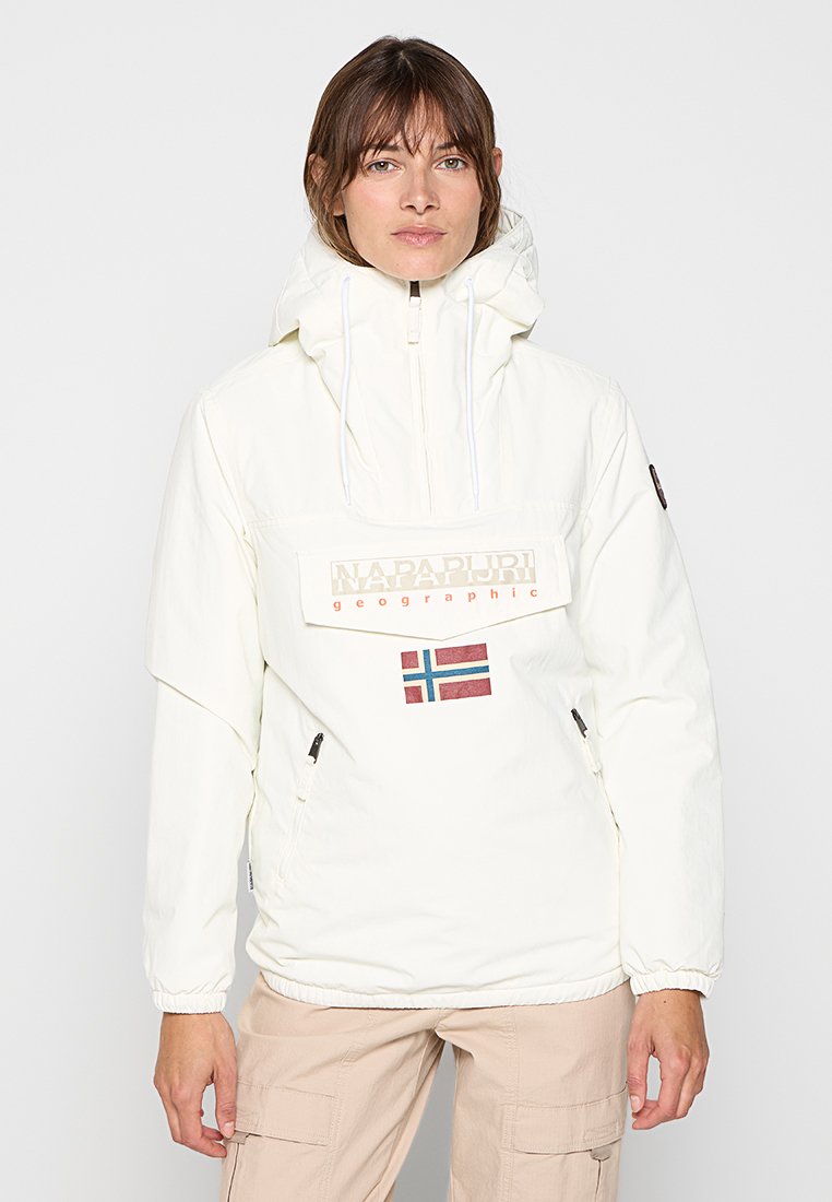 Junge Frau, die eine weiße Napapijri Kapuzenjacke mit einer norwegischen Flagge und beigefarbenen Cargo-Hosen trägt, vor einem schlichten Hintergrund steht.
