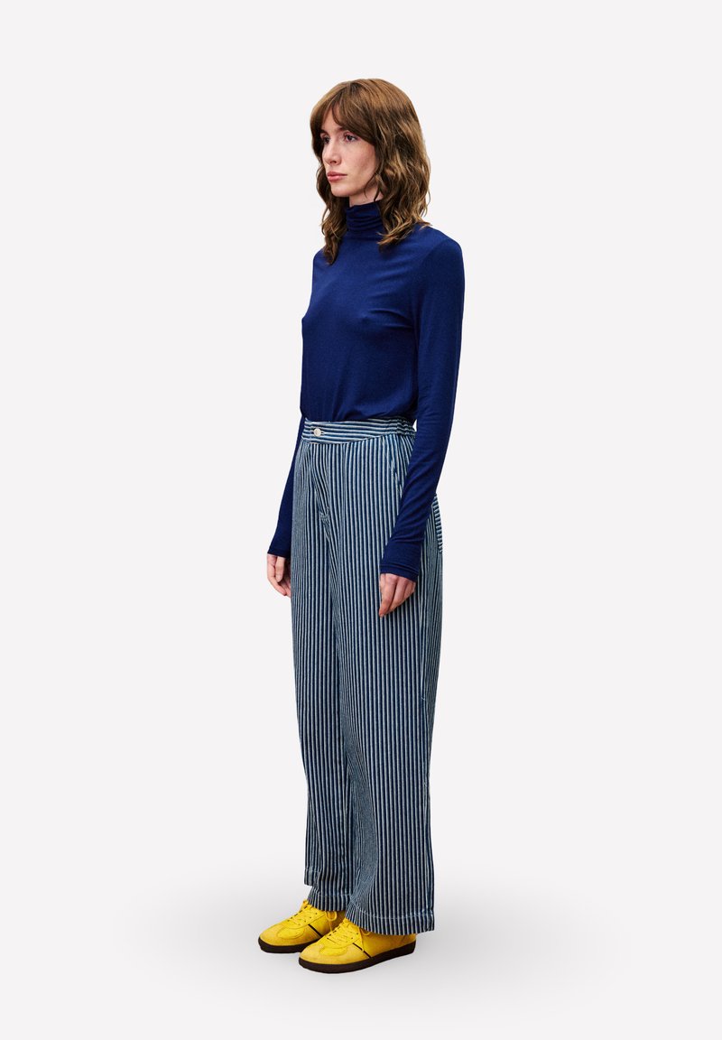 Frau in blauem Rollkragenpullover, hochtaillierten blau-weiß gestreiften Hosen und gelben Sneakers, die im Profil vor weißem Hintergrund steht.