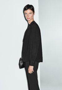 Blusa nera con una vestibilità rilassata, caratterizzata da un collo alto e maniche lunghe. Abbinata a pantaloni neri su misura e una piccola borsa nera.