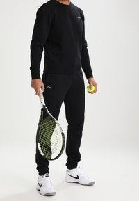 Sudadera negra y pantalones jogger de algodón con un acabado texturizado. La persona sostiene una raqueta de tenis y una pelota amarilla.