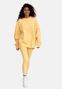 Żółta, przesadnie duża bluza z długim rękawem, zestawiona z żółtymi legginsami i białymi trampkami. Materiał wydaje się być miękki, o gładkiej teksturze.