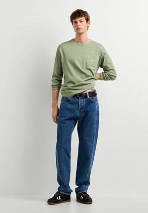 Un jeune homme se tient contre un mur blanc, portant une chemise verte à manches longues, un jean bleu avec une ceinture marron et des baskets noires.