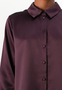 Dark purple button-up shirt with a collared neckline, smooth, shiny texture, and five front buttons. Long sleeves and minimal detailing.