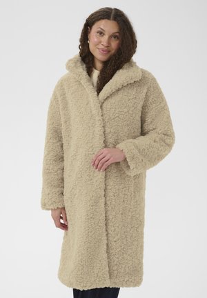 Kvinde med krøllet hår iført en lang beige teddyfrakke med krave, stående mod en ensfarvet hvid baggrund.