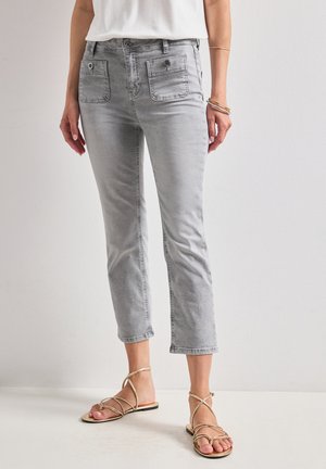 Femme portant un jean gris clair court avec poches à rabats à l'avant et des sandales plates dorées à brides, debout contre un mur blanc uni.