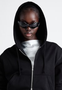 Felpa nera con zip, cappuccio regolabile e tasche frontali, abbinata a occhiali da sole neri affilati e angolari e a una maglietta grigia chiara.