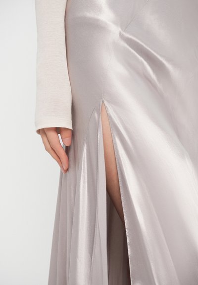 Χέρι που ακουμπά δίπλα σε γυαλιστερή ασημένια σατέν φούστα με σκίσιμο μέχρι το μηρό, δείχνοντας απαλή υφή υφάσματος και απαλό φυσικό φως.