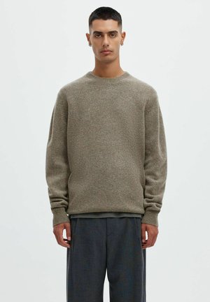 Giovane uomo con capelli corti e scuri che indossa un maglione grigio girocollo e pantaloni larghi grigio scuro, in piedi davanti a uno sfondo chiaro e semplice.