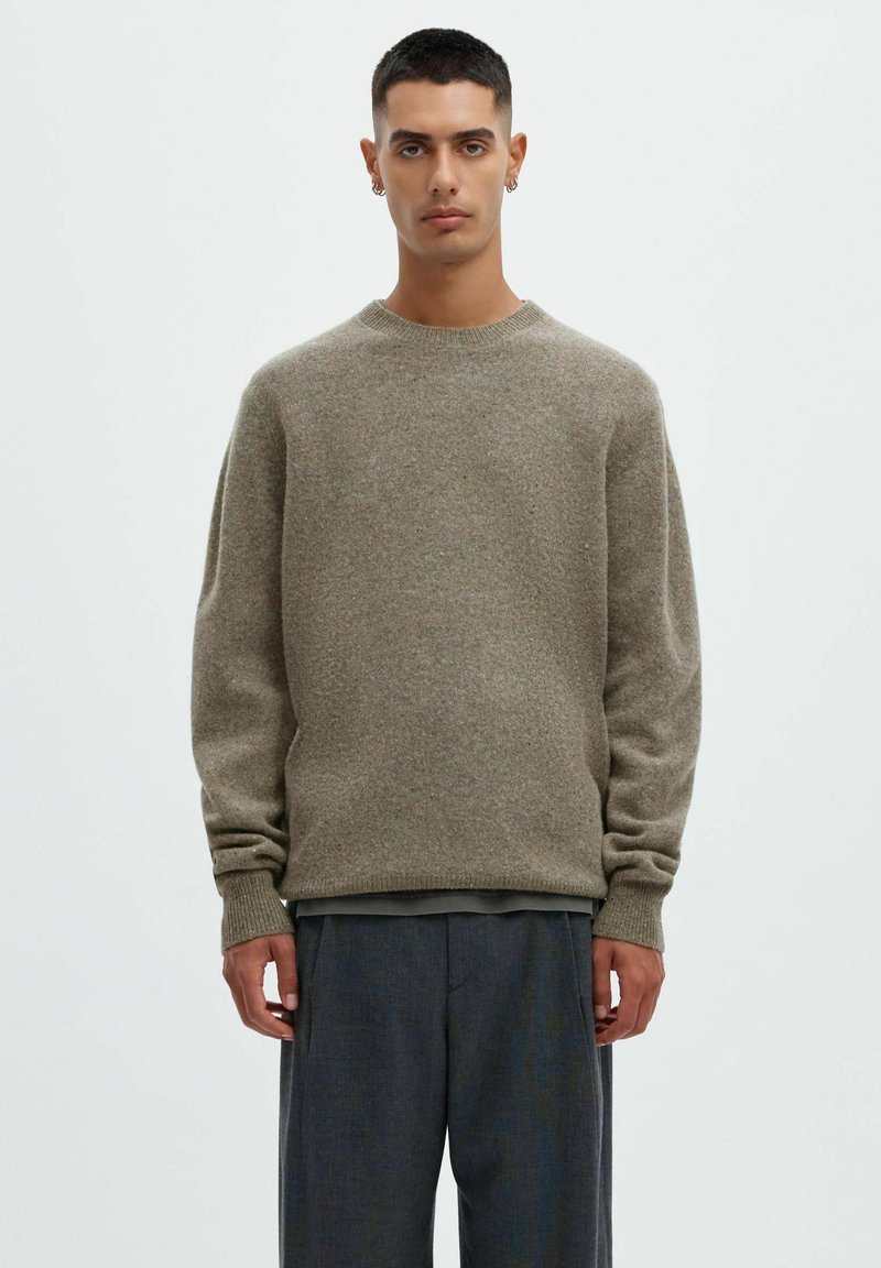 Jeune homme aux cheveux courts et foncés portant un pull gris à col rond et un pantalon ample gris foncé, debout devant un fond clair uni.