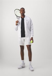 Weiße Sportjacke über einem schwarzen Shirt, weiße Shorts und weiße Nike-Sneaker. Hält einen Tennisschläger und einen gelben Tennisball.