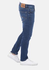 Jeans in denim blu con una vestibilità slim, caratterizzati da una patta in pelle marrone in vita e da cuciture discreti sulle tasche posteriori. Indossati con sneakers bianche.