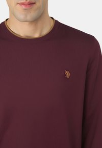 Uomo che indossa una maglia a maniche lunghe bordeaux a coste con un piccolo emblema ricamato in oro sul petto sinistro e una rifinitura marrone sul collo.