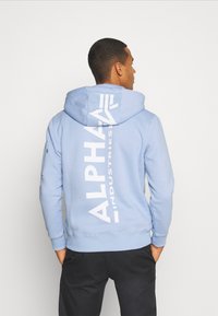 Felpa blu chiaro con cappuccio regolabile. Grande grafica bianca "ALPHA INDUSTRIES" sul retro, con una tasca laterale con zip.