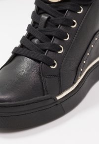 Zapatilla alta de cuero negro con superficie texturizada, suela de goma contrastante y ojales de metal. Presenta cordones negros gruesos y tachuelas.