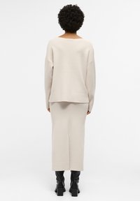 Robe en maille beige, ajustée, longueur genou, manches longues et large encolure, associée à des bottes noires à talons hauteur genou. Texture lisse.