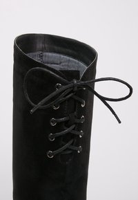 Bottine en  daim noir avec un bout rond, dotée d'œillets métalliques et de lacets. La doublure est grise et le dessus présente un léger pli.