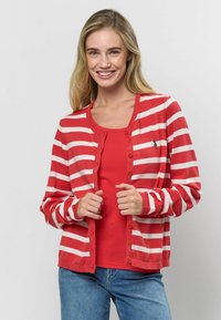 Červený a biely pruhovaný kardigán s gombíkovým zapínaním a rebrovanými manžetami, nosený cez jednofarebný červený top, spolu s modrými džínsami.