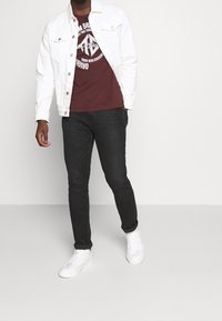 Chaqueta de mezclilla blanca, camiseta gráfica en color burdeos, pantalones vaqueros negros y zapatillas blancas. El conjunto presenta un diseño casual y texturas suaves.