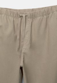 Pantaloni ușori bej, cu talie elastică și snur. Prezintă un design simplu și o textură netedă. Fără buzunare vizibile.