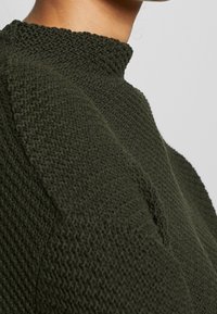 Sweter dzianinowy z wysokim kołnierzem, teksturowanym ciemnozielonym materiałem i charakterystycznym krojem ramion. Zbliżenie na materiał i kształt.