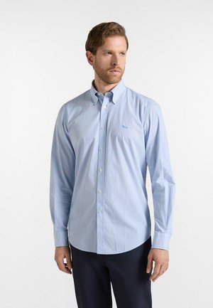 Hombre con camisa de manga larga azul claro abotonada y pantalones oscuros, de pie con una expresión neutral sobre un fondo blanco liso.
