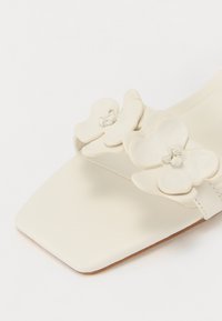 Krämfärgade sandaler med fyrkantig tå, med en blommig läderdetalj med lager av kronblad på remmen, slät textur, minimalistisk design.