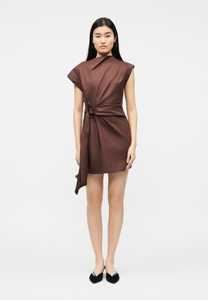 Naine seisab, kandes varrukateta pruuni minikleiti, millel on asümmeetriline drapeeringu detail, ning musti terava ninaga lameda tallaga kingi, taust on lihtne.