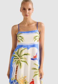 Νεαρή γυναίκα φορώντας φόρεμα με τροπικό μοτίβο σε μπλε και άσπρο χρώμα, με φοίνικες, πολύχρωμους παπαγάλους και λουλουδένια σχέδια, στέκεται μπροστά σε απλό φόντο.