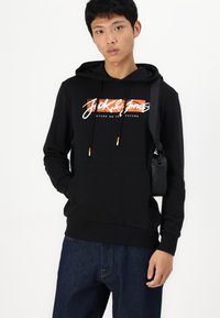 Sudadera negra con capucha que presenta un bolsillo frontal, cordones con acentos en naranja y un logo gráfico en blanco y naranja. Combinada con pantalones de mezclilla oscura.