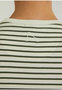 Rückansicht einer Person, die ein cremefarbenes und dunkelgrünes, horizontal gestreiftes Hemd trägt, mit einem aufgenähten Buchstaben "A" im oberen Rückenbereich.